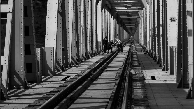 Melnbalta fotogrāfija ar dzelzceļa tilta metāla konstrukciju iekšskatu. Pa vidu redzamas sliedes, gar kurām vairāki cilvēki veic sliežu vai infrastruktūras uzturēšanas darbus.