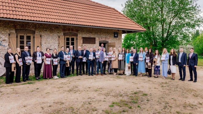 Grupas foto ārpusē pie mūra ēkas ar sarkanu dakstiņu jumtu, kur rindā stāv aptuveni trīsdesmit cilvēku, katrs rokās turot diplomu vai apbalvojumu, kas norāda uz publisku apbalvošanas vai pasākuma ceremoniju.