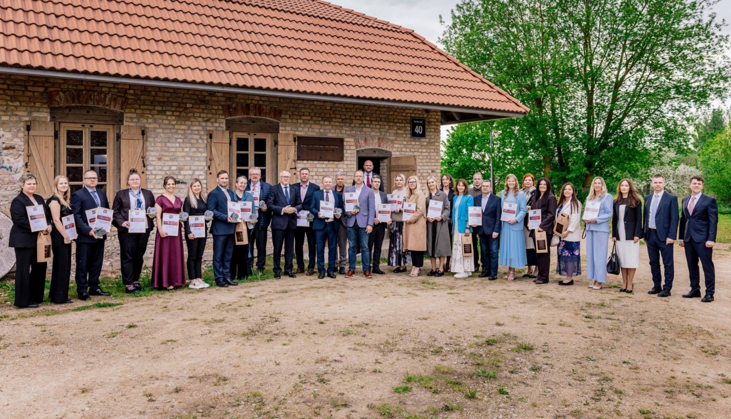 Grupas foto ārpusē pie mūra ēkas ar sarkanu dakstiņu jumtu, kur rindā stāv aptuveni trīsdesmit cilvēku, katrs rokās turot diplomu vai apbalvojumu, kas norāda uz publisku apbalvošanas vai pasākuma ceremoniju.