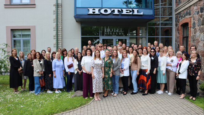 Cilvēki (dalībnieki), māja, zāliens, bruģēts ceļš, uzraksts "Hotel"