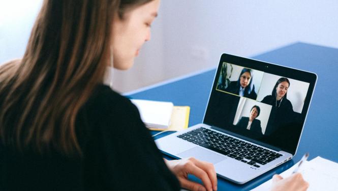 Sieviete sēž pie gada uz kura novietots dators un sarunājas ar cilvēkiem tiešsaistē, izmantojot video konferences iespējas. 