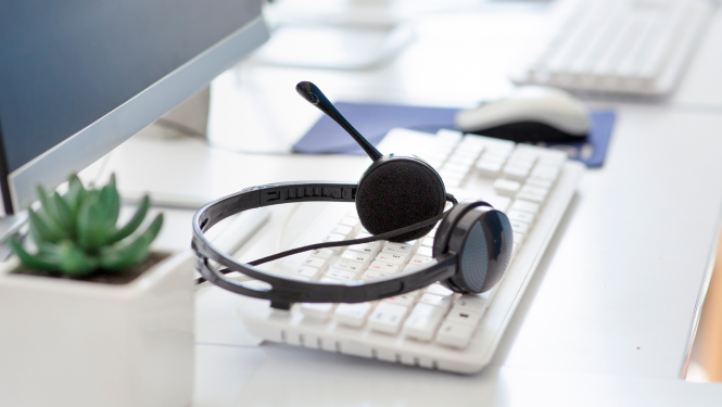 Austiņas ar mikrofonu novietotas virs baltas klaviatūras, fonā datorpele, monitors, telpaugs