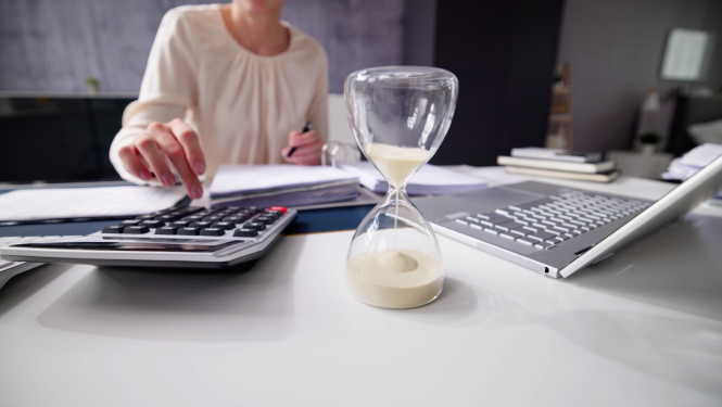 Sieviete aprēķina datus uz kalkulatora ar labo roku, pretējā pusē dators, priekšā viņai smilšu pulkstenis 