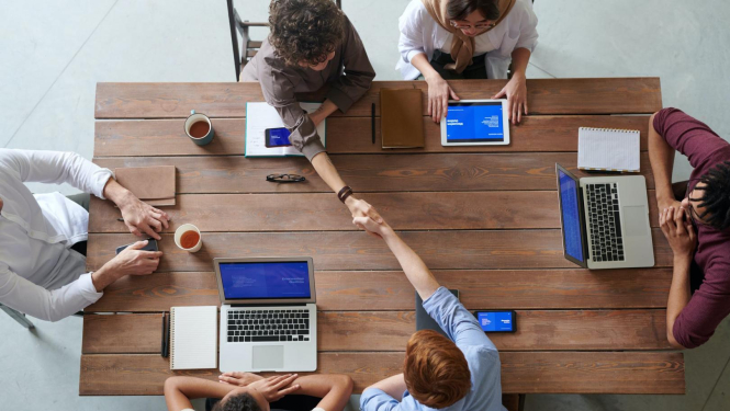 Pie darba galda sēž seši cilvēki un strādā, sarunājas, pielieto datortehniku