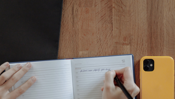 Cilvēks kladē pieraksta svarīgāko, blakus kladei novietots telefons