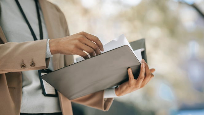 Persona gaišā žaketē un baltā kreklā tur atvērtu mapi, kurā ievieto vai izņem dokumentus. Fons ir izpludināts, ar dabisku apgaismojumu.